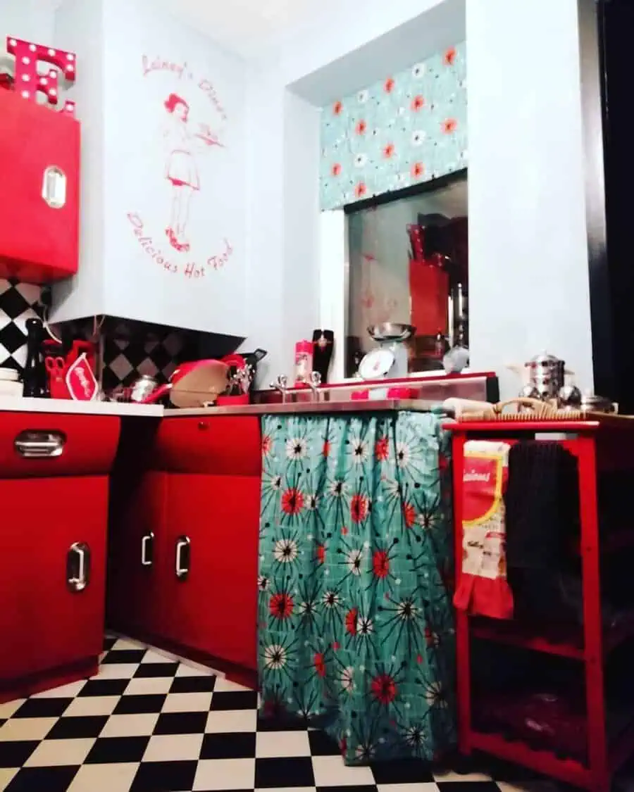 1950s kitchen with red cabinets, checkered floor, and floral-patterned curtain accents.