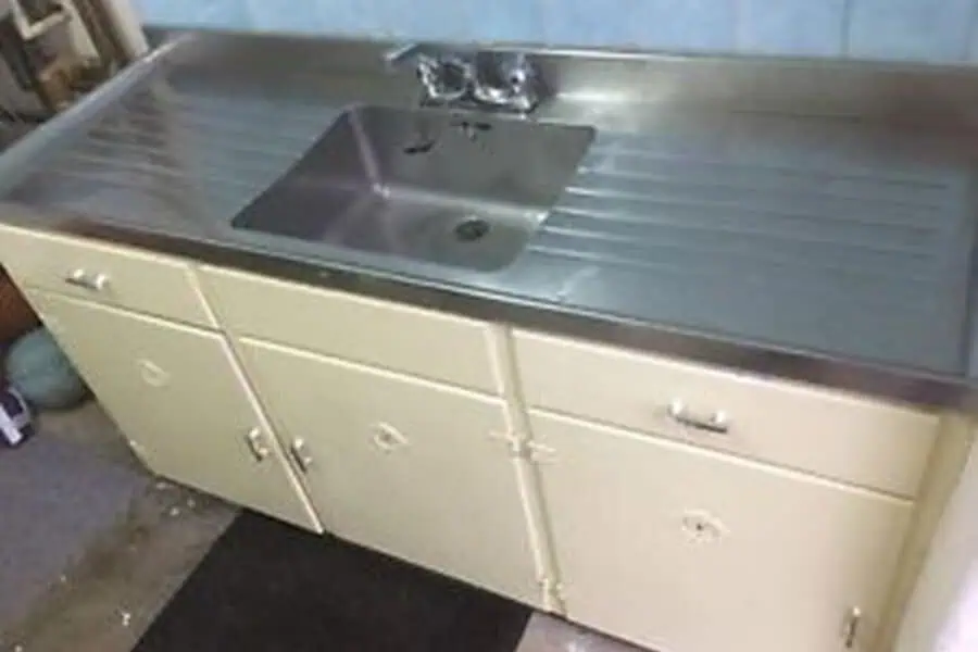 1950s kitchen sink with stainless steel countertop and cream-colored metal cabinets.