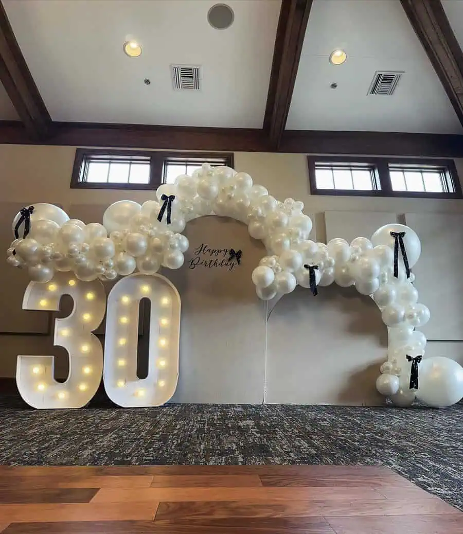 Elegant white balloon arch with illuminated "30" and Happy Birthday backdrop.