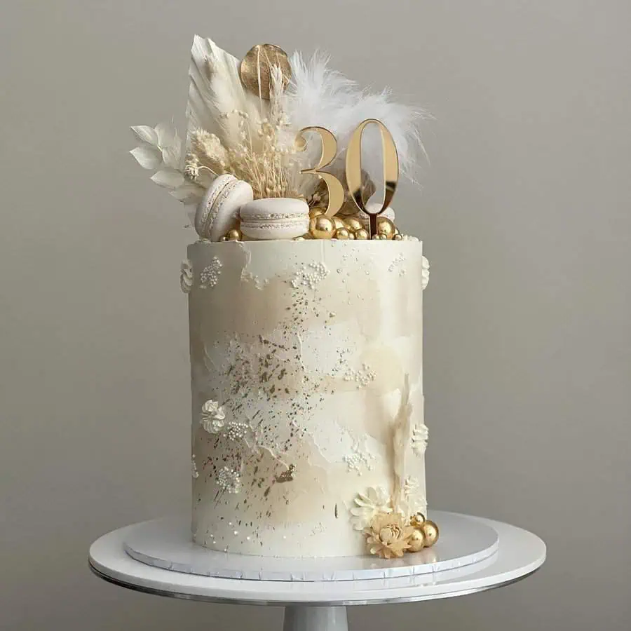 Elegant beige and gold 30th birthday cake with macarons, feathers, and metallic accents.