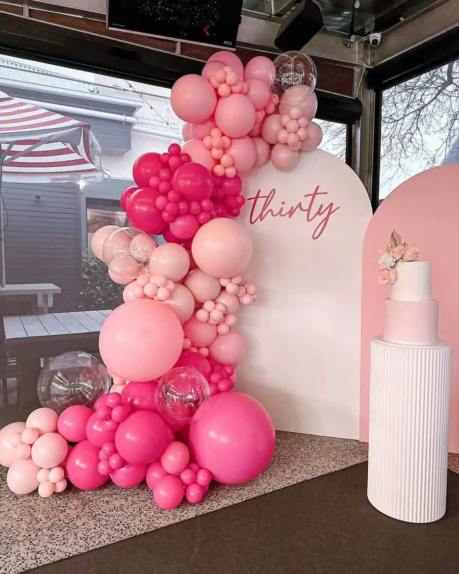 Pink balloon garland with "thirty" sign and minimalist cake for a 30th birthday celebration.