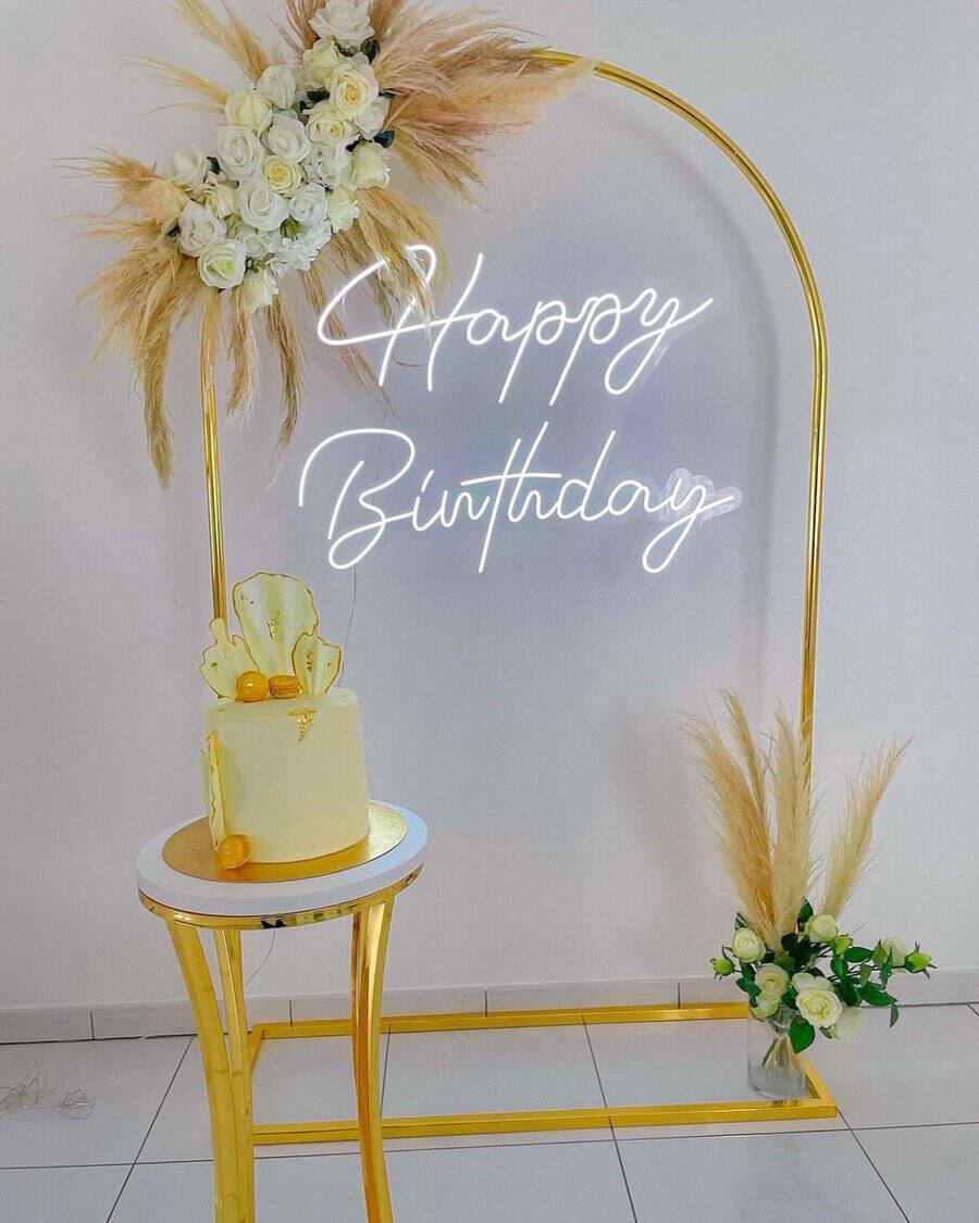 Minimalist birthday party with gold accents, pampas grass, white roses, and a neon "Happy Birthday" sign.
