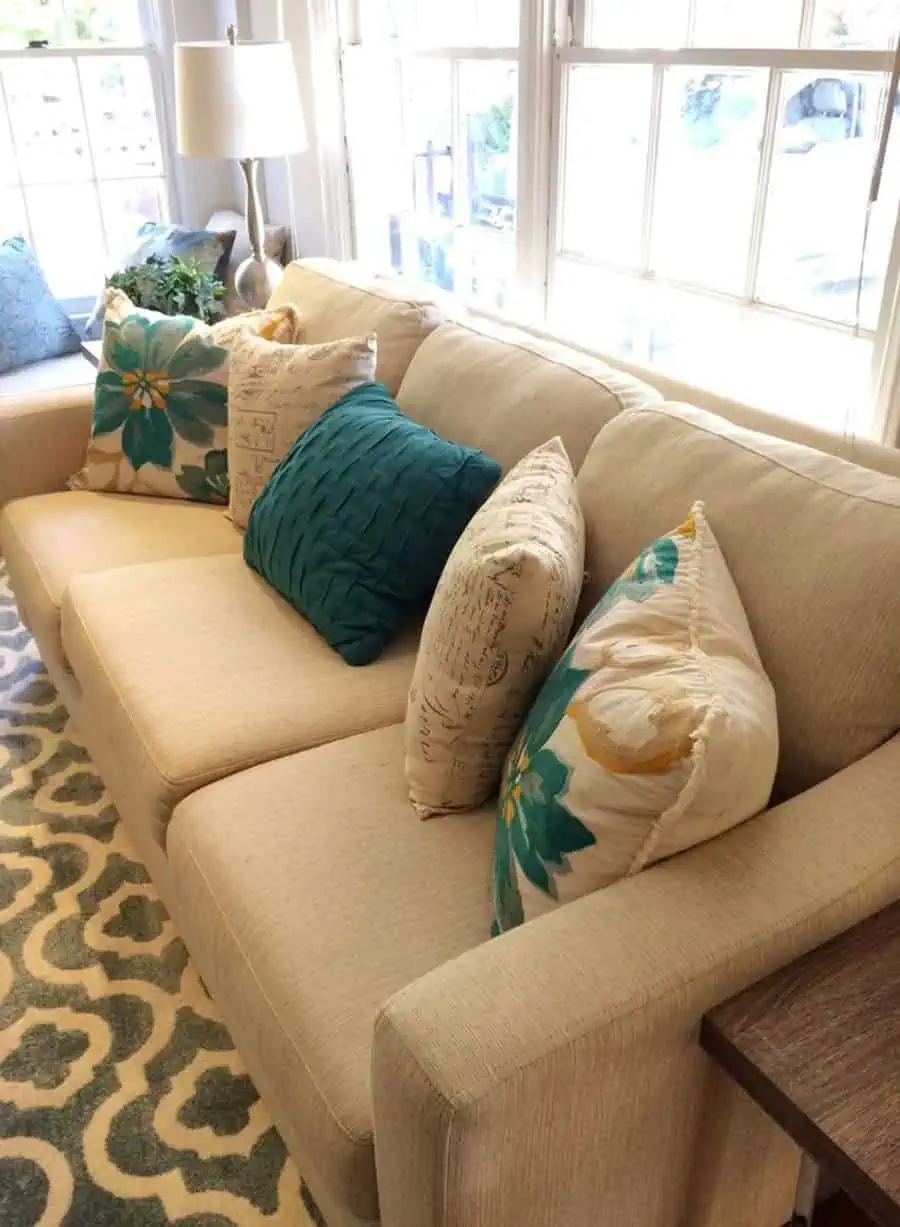 Beige sofa with teal and floral pillows near large windows and a patterned rug.