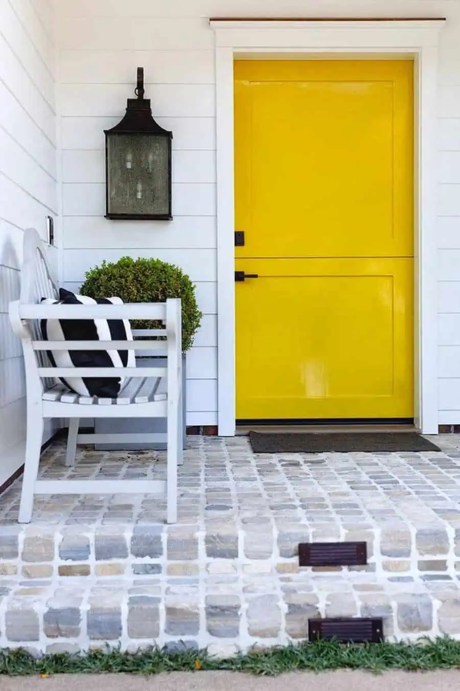 Bright yellow Dutch door with black hardware, white siding, and a brick porch with seating.