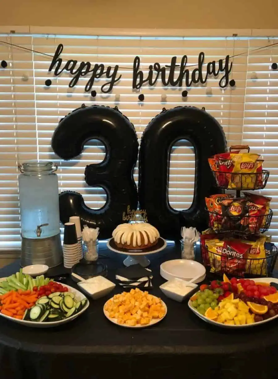 Budget-friendly 30th birthday table with snacks, fruit, cake, and black balloon decor.