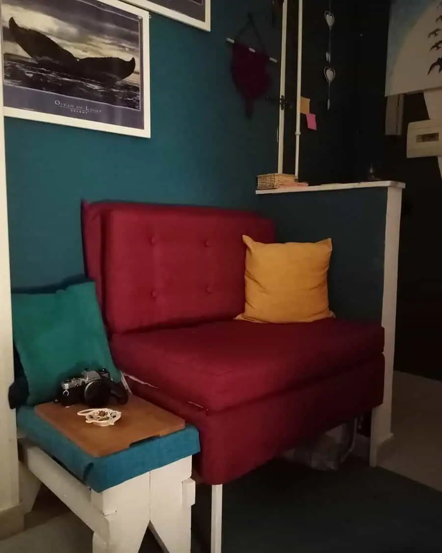 Cozy corner with burgundy chair, teal and mustard cushions, and teal walls with framed artwork.