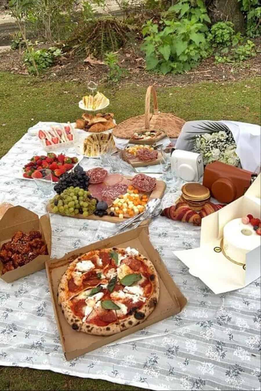 Outdoor picnic spread with pizza, sandwiches, fruits, cheese, and desserts on a blanket.