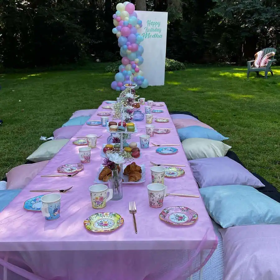 Pastel-themed outdoor birthday table with floral plates, pillows, and a balloon backdrop.