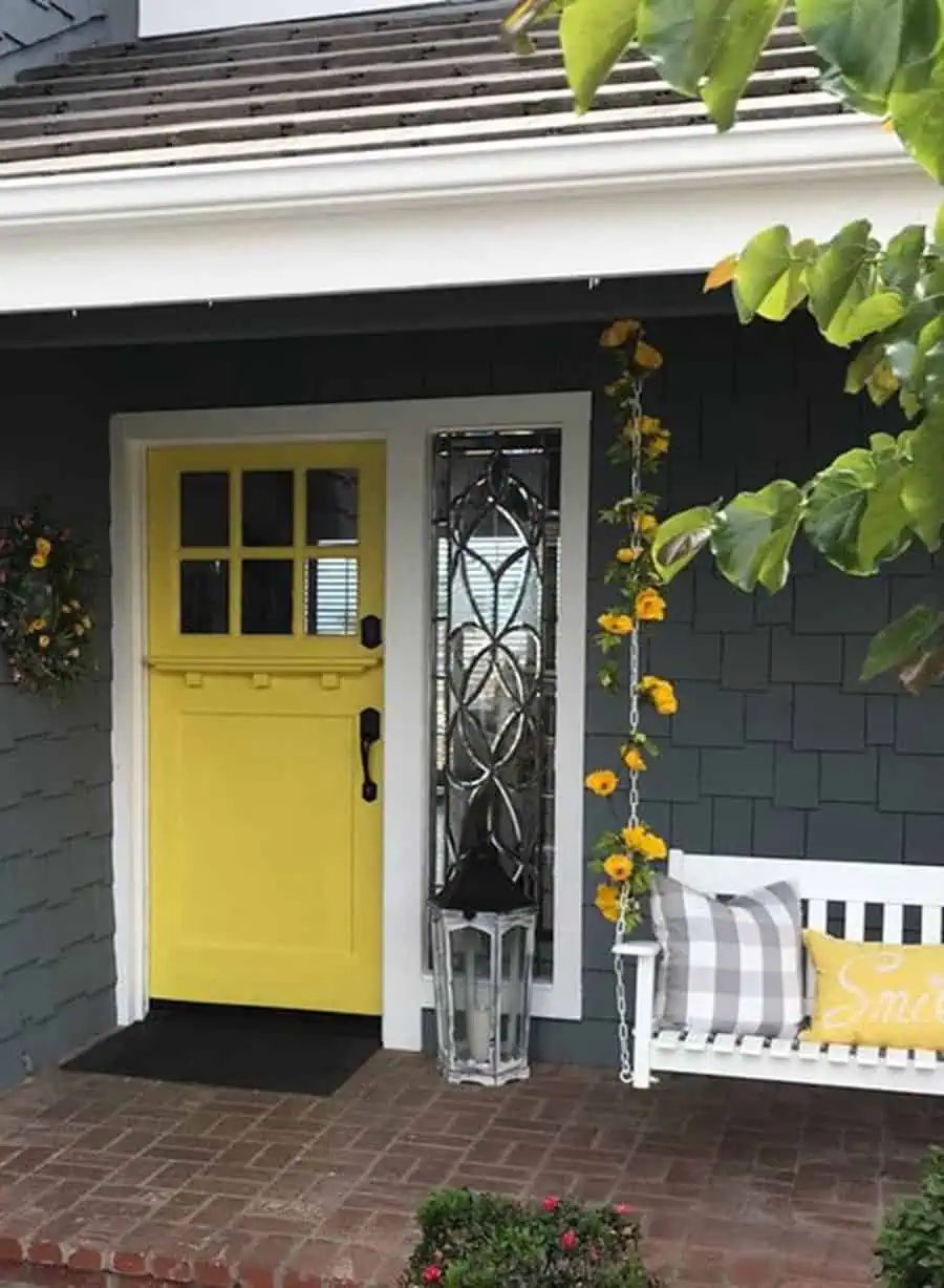 Yellow Dutch door with decorative glass side panel, brick porch, and cozy swing adorned with pillows.