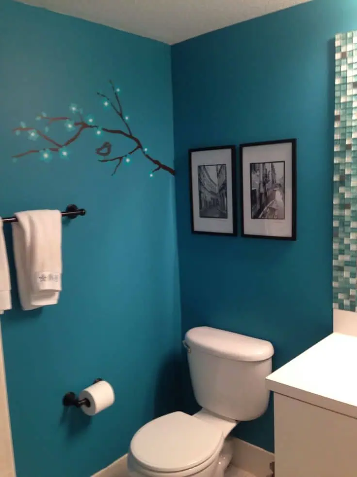 Teal bathroom with branch wall decal, black-framed art, and white fixtures.