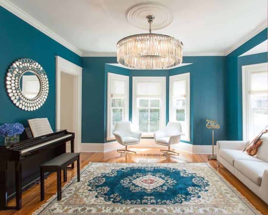 Teal living room with white chairs, crystal chandelier, piano, and an ornate teal area rug.