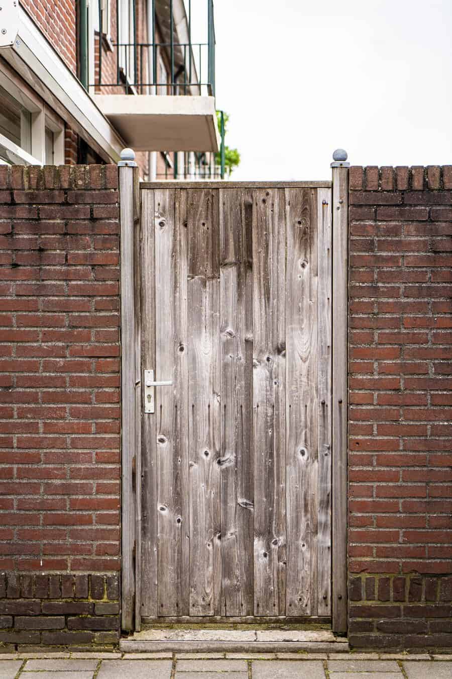 Weathered wooden gate with a white handle, framed by red brick walls in an urban setting.