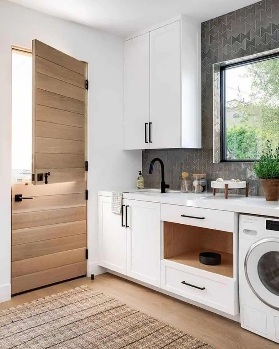 Modern laundry room with wooden Dutch door, white cabinets, black faucet, and large window.