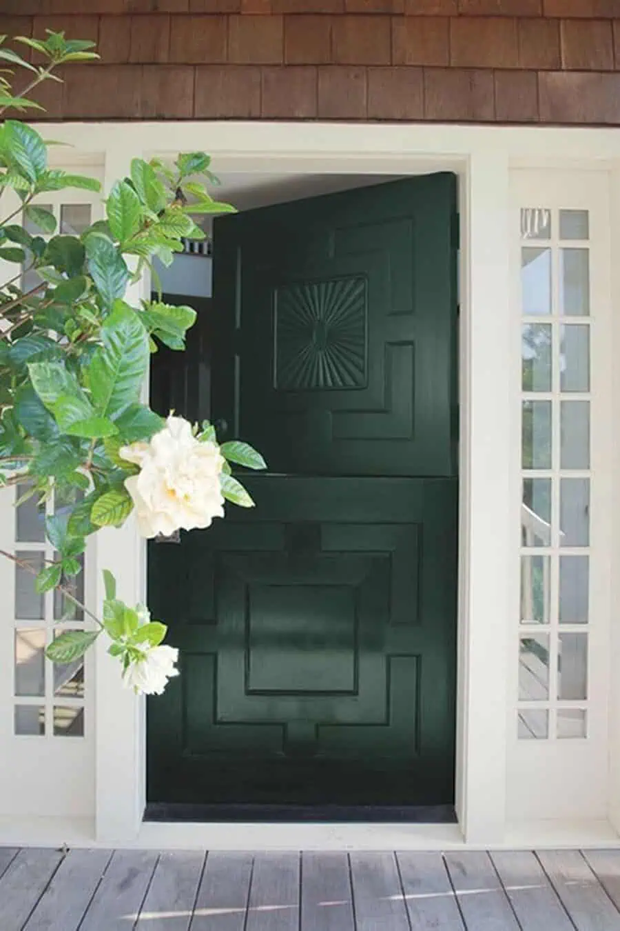 Dark green Dutch door with intricate design surrounded by white trim and blooming flowers.