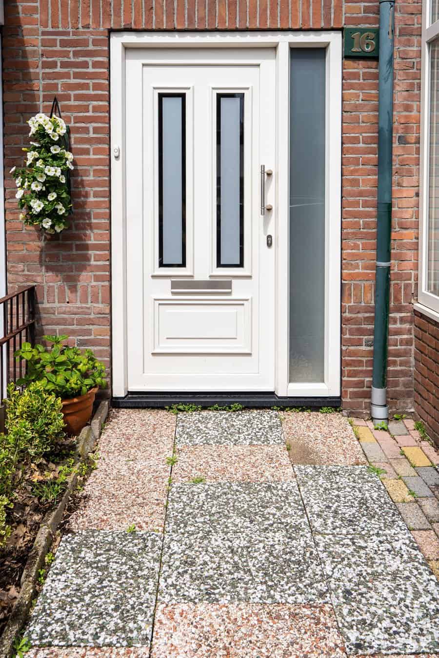 White front door with glass panels, brick wall, and potted plants on a tiled pathway.