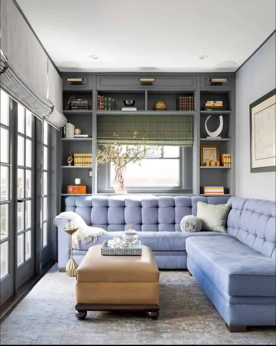 Modern den with built-in shelves, blue sectional sofa, leather ottoman, and natural light.