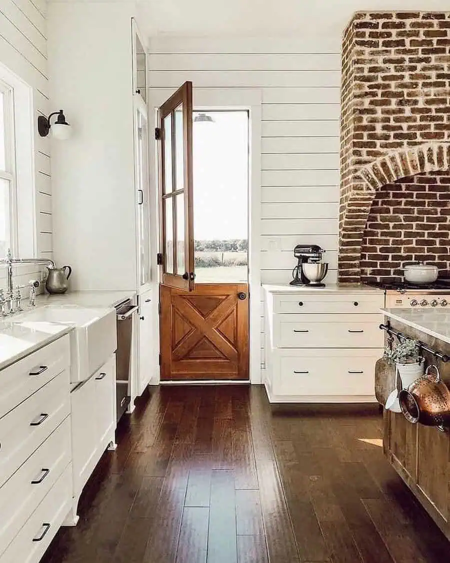 Farmhouse kitchen with wooden Dutch door, brick fireplace, and white cabinetry on hardwood floors.