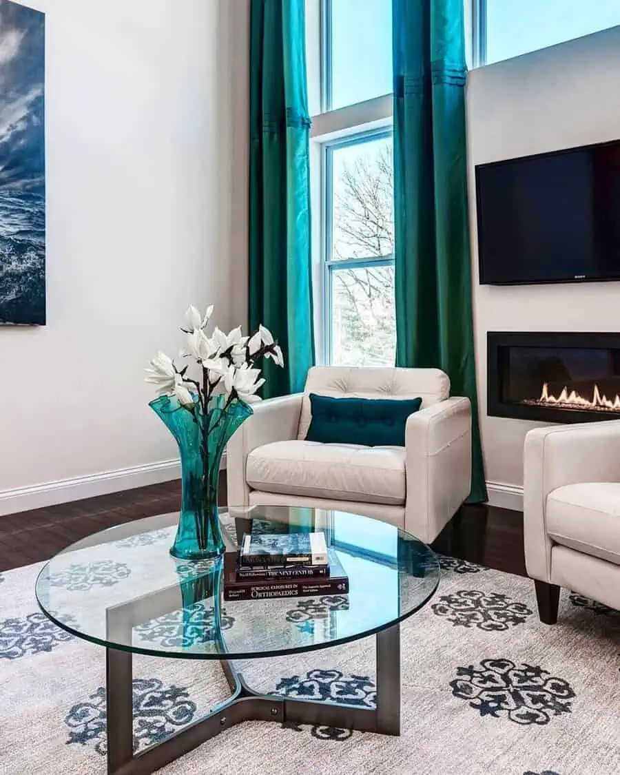 Teal living room with floor-length curtains, a glass coffee table, and cozy seating by the fireplace.