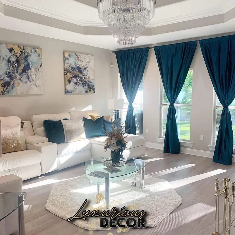 Luxurious teal living room featuring floor-length curtains, elegant white seating, and a sparkling chandelier.