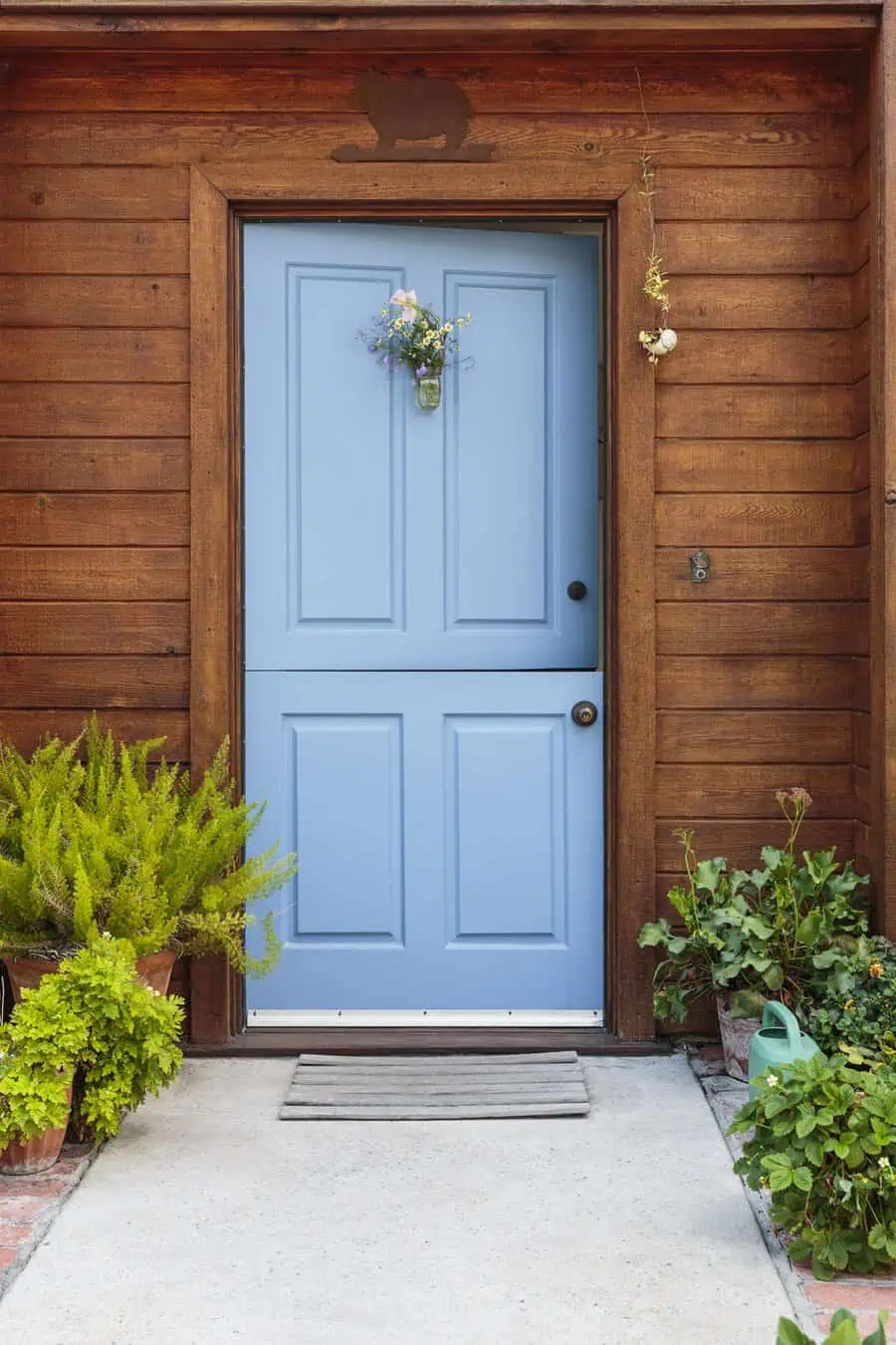 Blue Dutch door with a floral wreath, wooden siding, and potted greenery on a front porch.