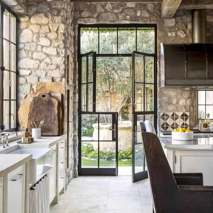 Rustic kitchen with stone walls, black-framed glass doors, and a view of a lush garden courtyard.