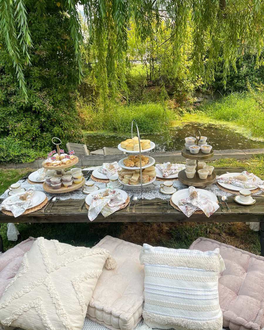 Elegant outdoor tea party setup with pastries, fine china, and cushions under a willow tree.