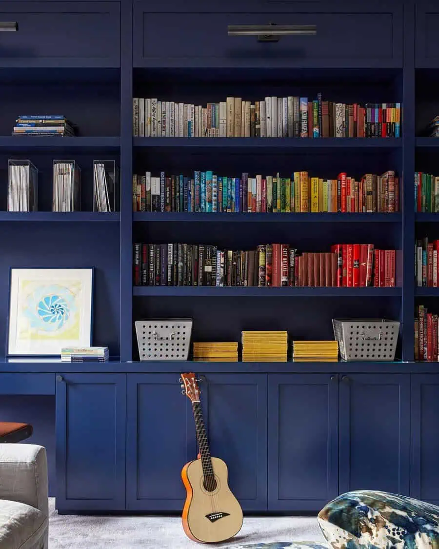 Blue built-in bookshelves with colorful books, decorative baskets, and a guitar in a cozy den.