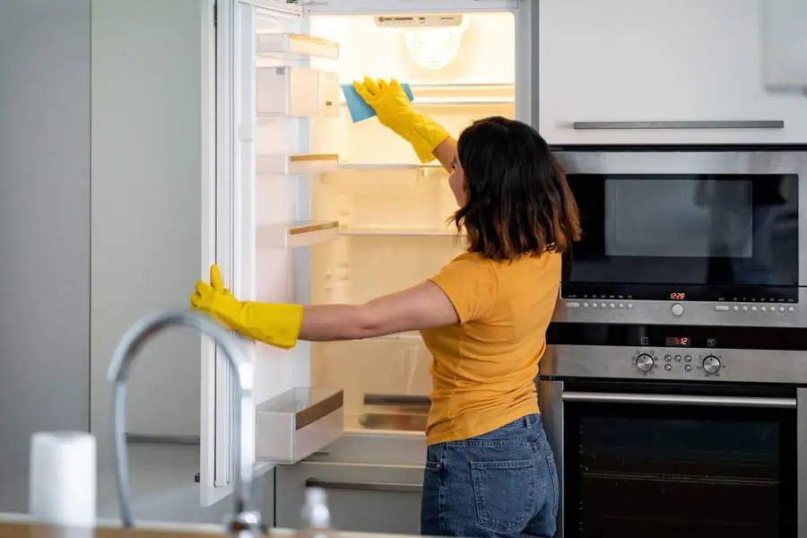 Woman cleaning an empty refrigerator with gloves and a cloth in a modern kitchen.