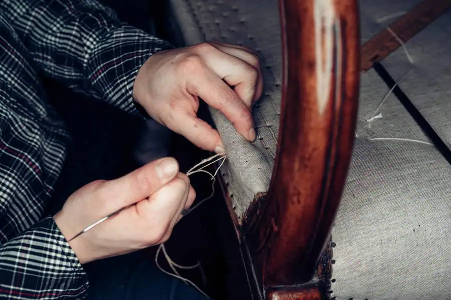 Hands sewing fabric onto a wooden chair frame with a needle and thread during reupholstery.