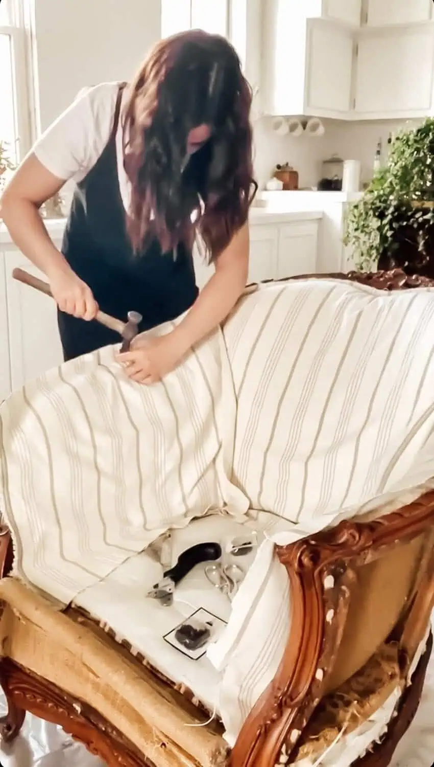 Woman using a hammer to attach striped fabric to a vintage chair during reupholstery.