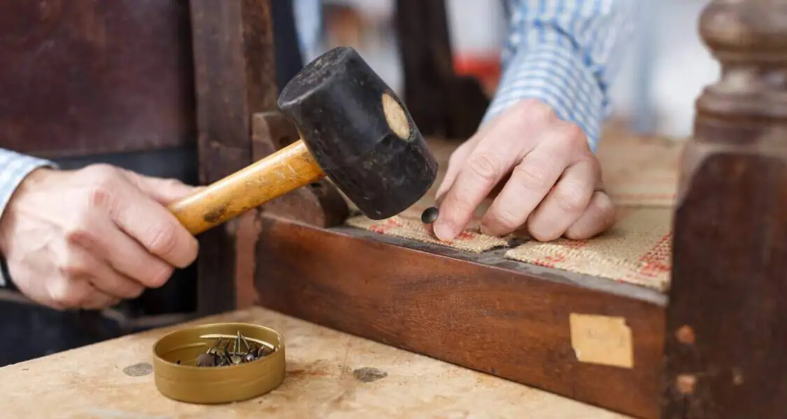 Person using a mallet to hammer decorative tacks into a chair frame during reupholstery.