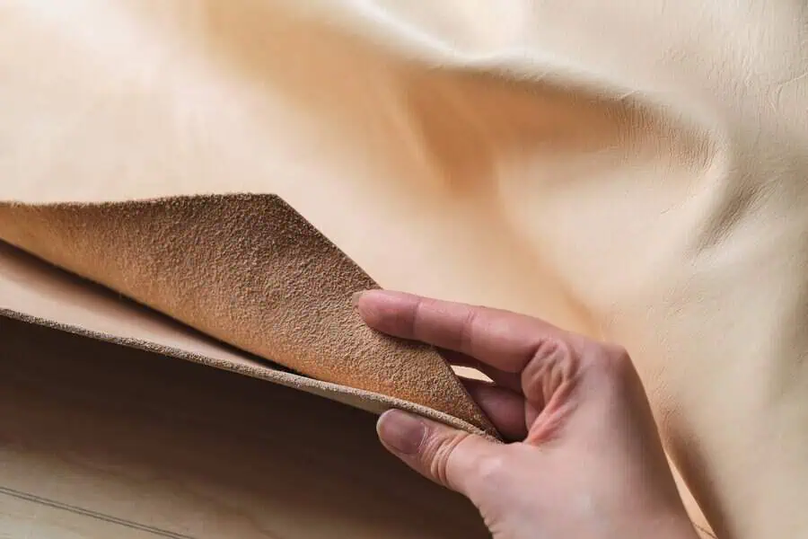 Close-up of a hand holding beige leather fabric, showing its smooth and textured sides.