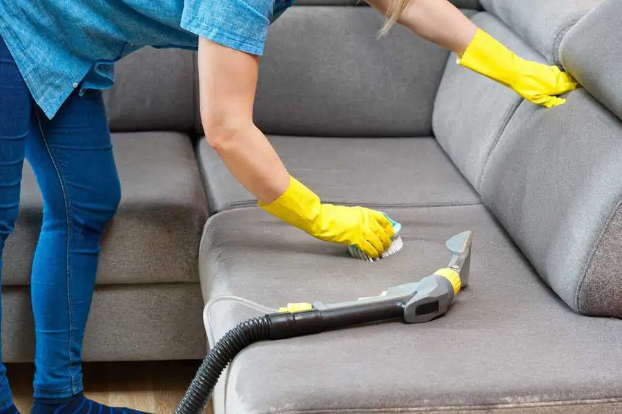 Person cleaning a grey sofa using a brush and vacuum cleaner while wearing yellow gloves.