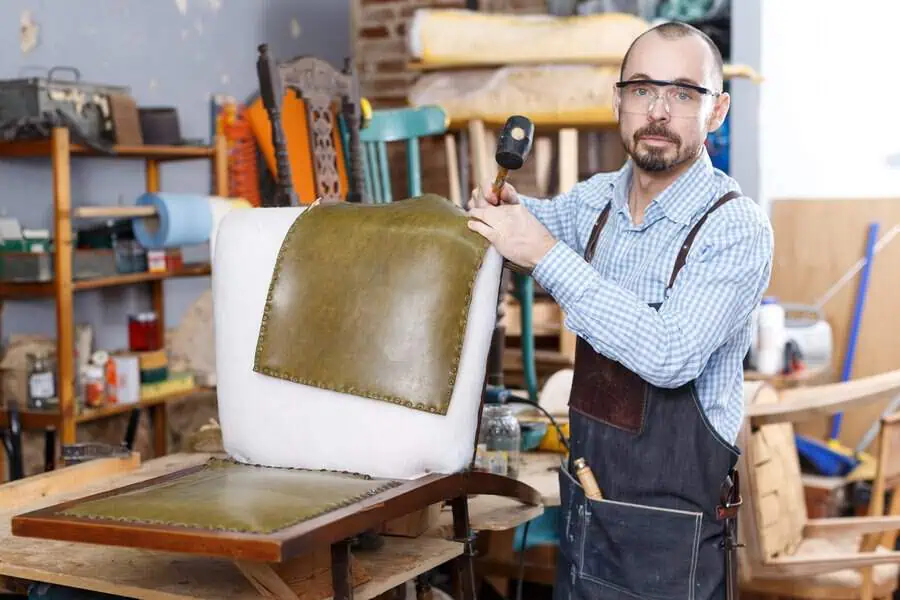 Man reupholstering a chair with green leather in a workshop wearing safety glasses and apron.