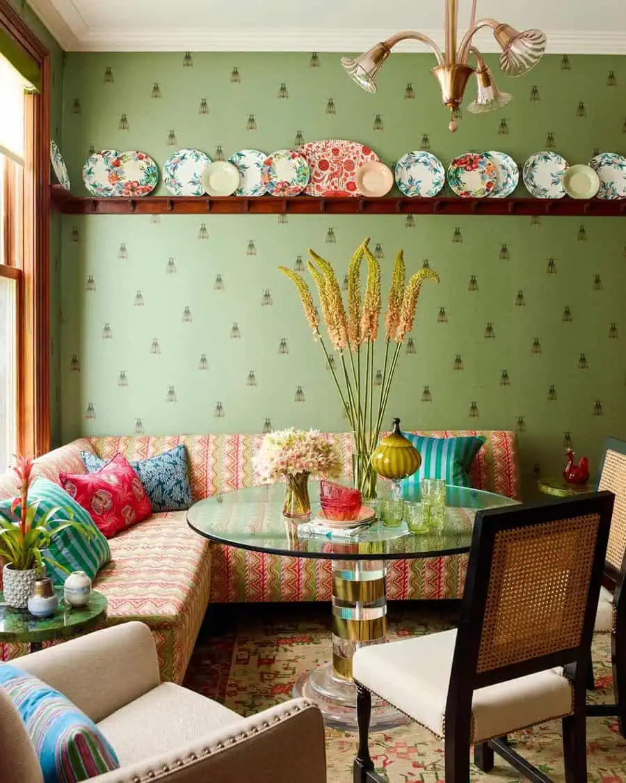 Eclectic dining area with patterned sofa, green wallpaper, colorful plates, and glass table.