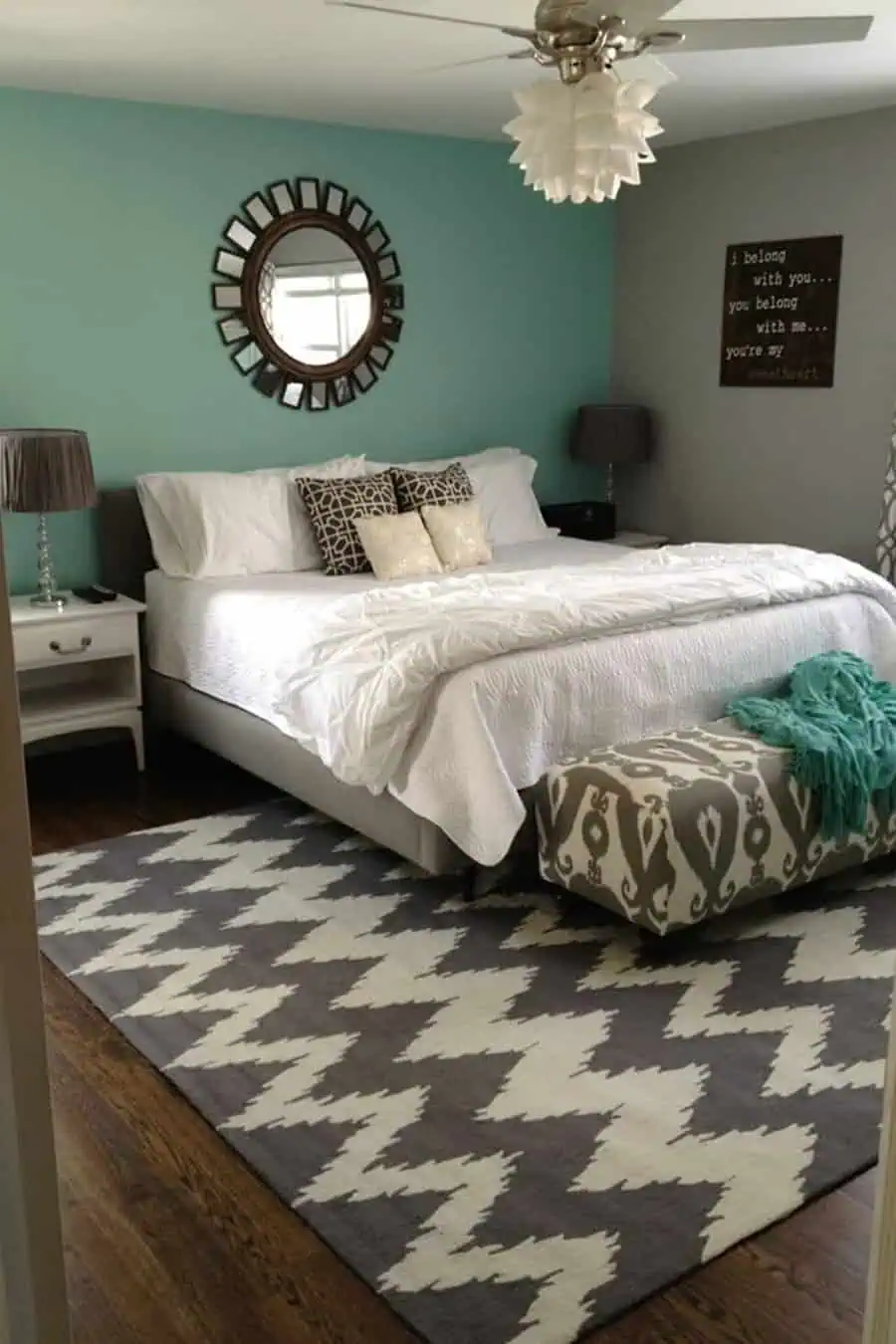 Bedroom with teal and light gray walls, white bedding, patterned bench, and chevron area rug.