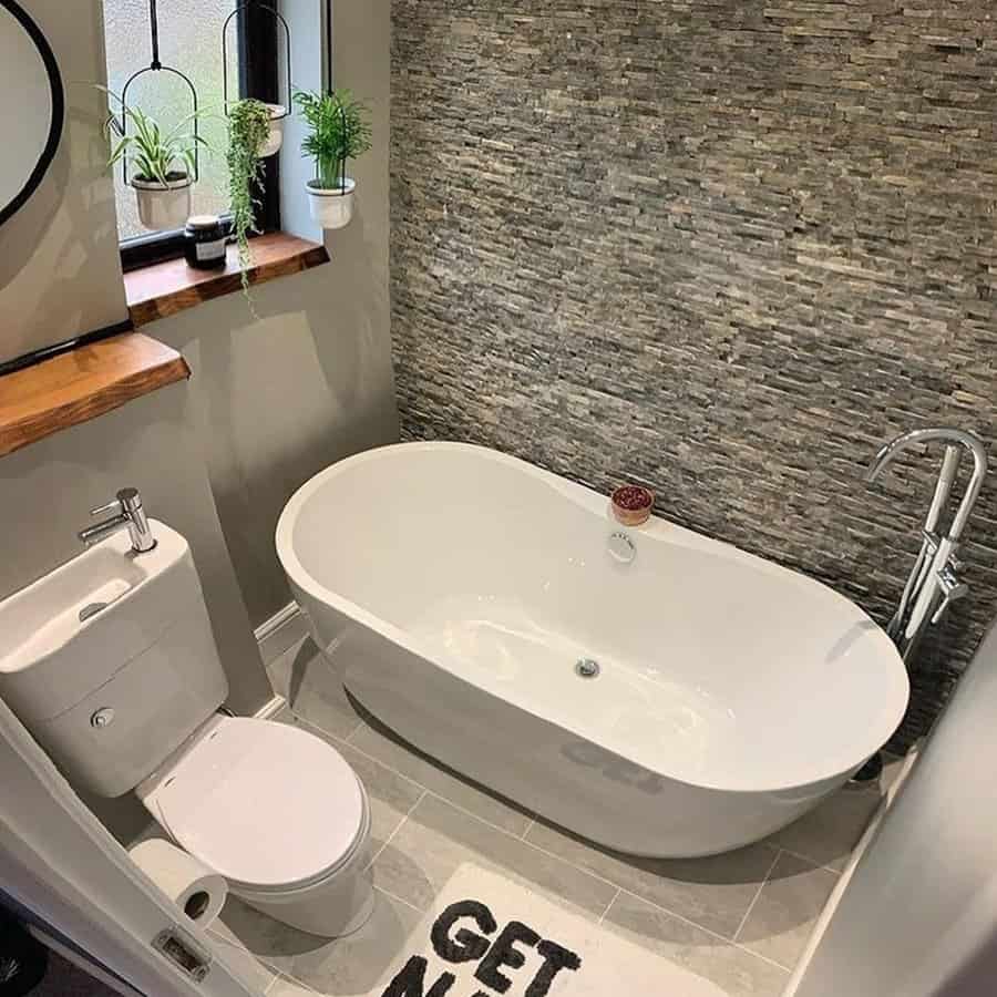 Small luxury bathroom with stone accent wall, freestanding tub, wood shelf, hanging plants, and luxe colors.