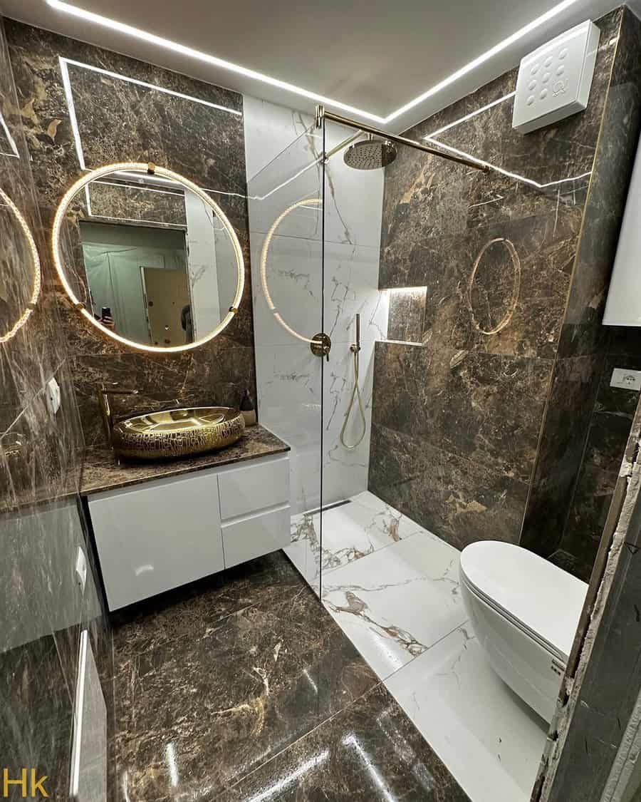 Small luxury bathroom with marble walls, gold accents, LED mirrors, and a glass shower enclosure.