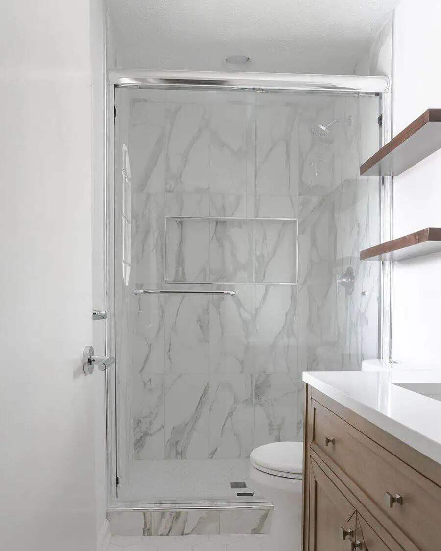 Small luxury bathroom with marble tiles, glass shower door, wooden vanity, and floating shelves.