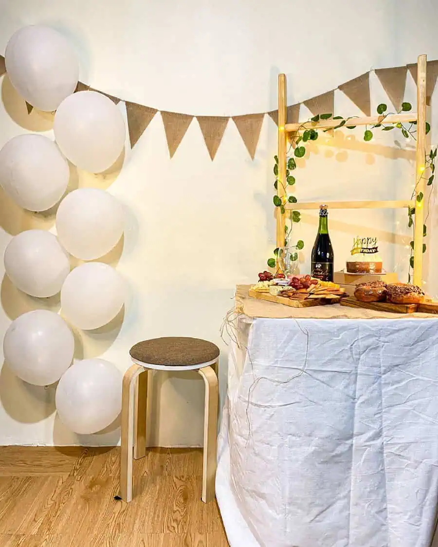 Minimalist birthday setup with white balloons, rustic decor, and a dessert table with cake and snacks.