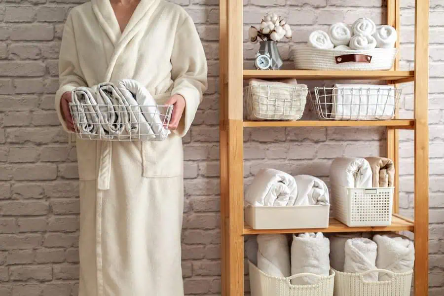 Person in a robe holding folded towels next to a wooden shelf with neatly organized linens.