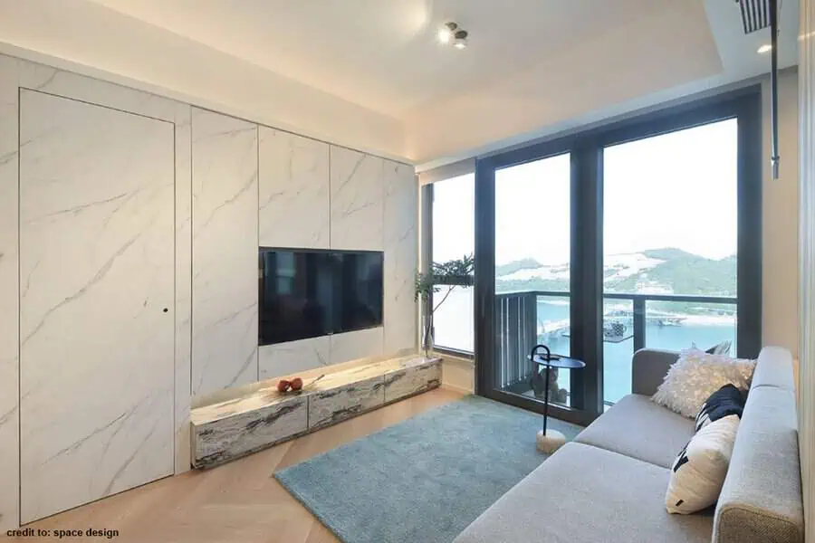 Modern living room with marble walls, wall-mounted TV, large windows, and a cozy grey sofa.