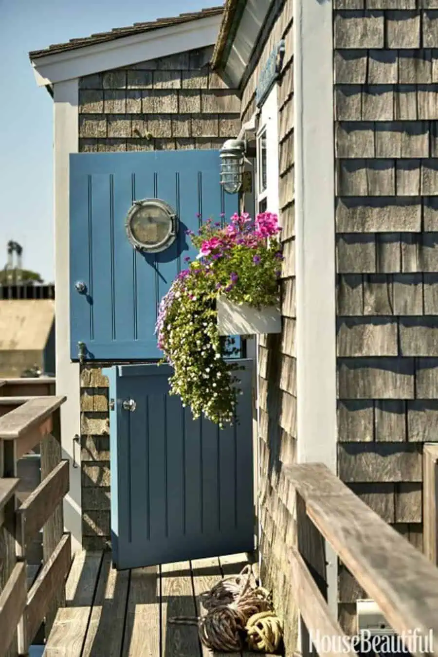 Blue Dutch door with porthole window on a rustic balcony with flowers and nautical decor.