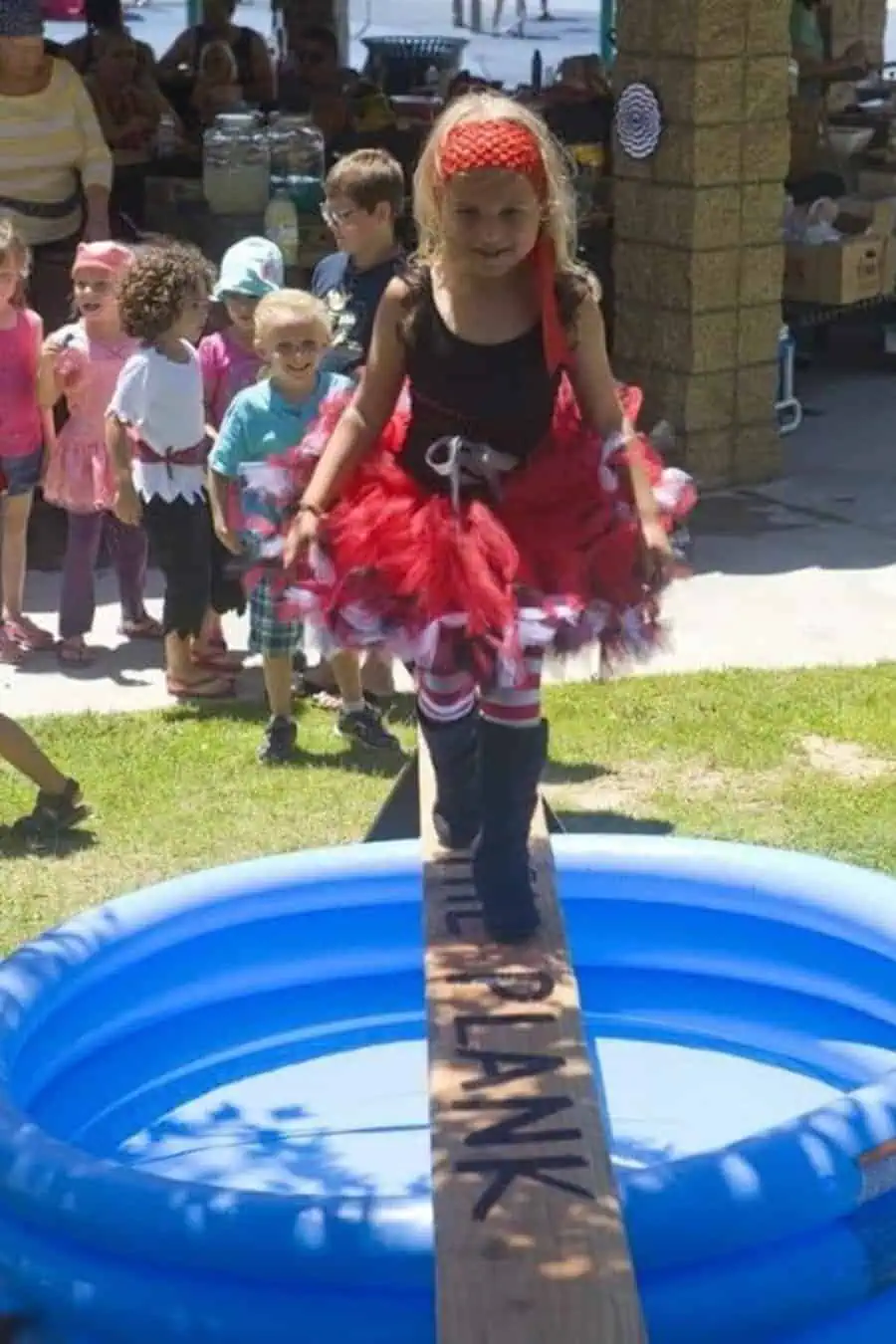 Pirate-themed outdoor birthday game with kids walking the plank over a kiddie pool.