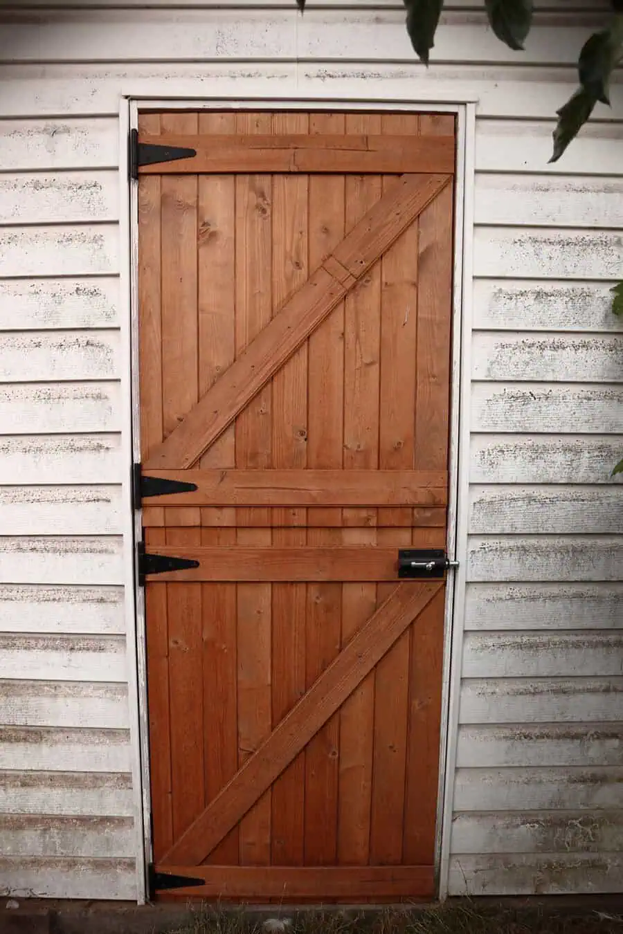 Rustic wooden Dutch door with diagonal planks, black hinges, and white siding background.