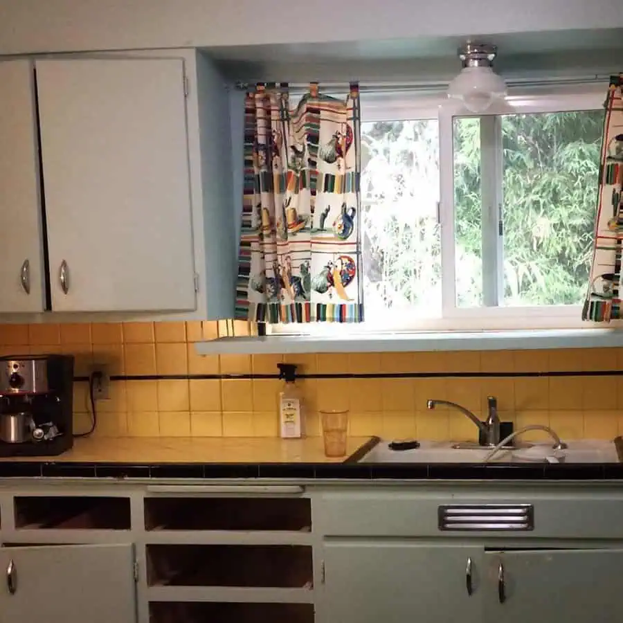 Retro 1950s kitchen with rooster-print curtains, pastel blue cabinets, and yellow tiles.