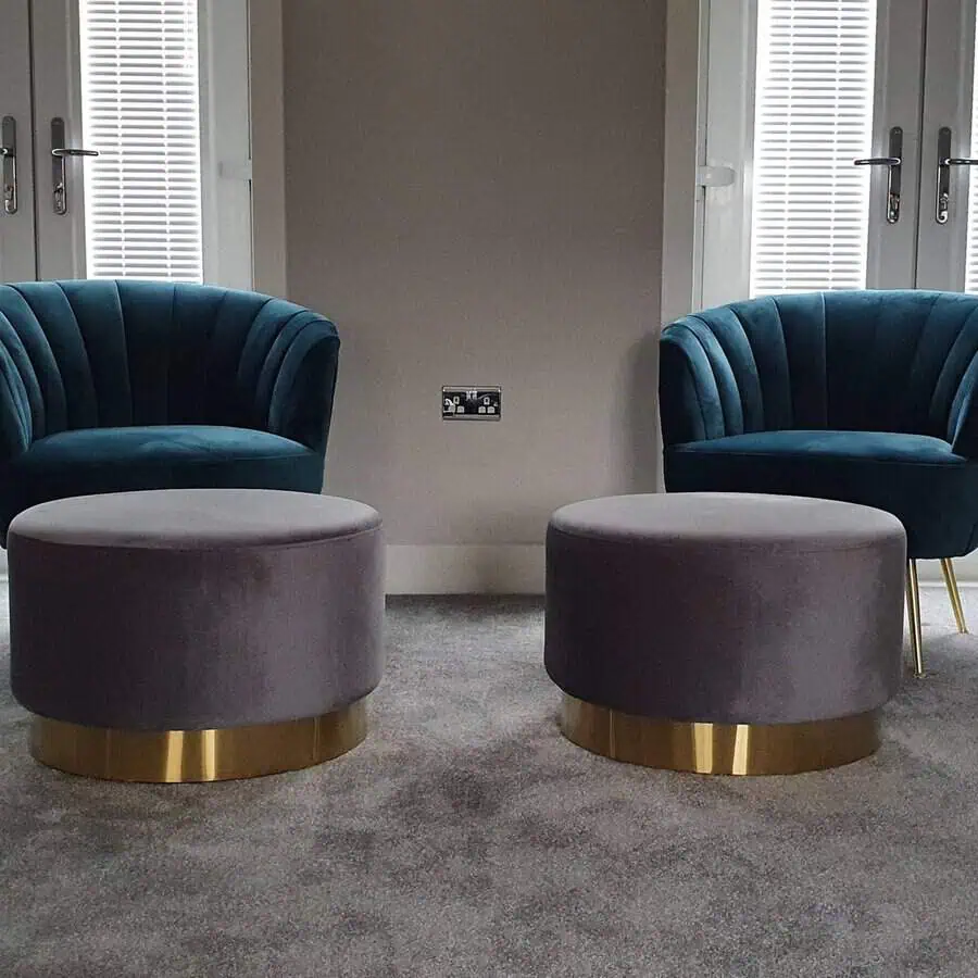 Teal living room with velvet chairs, gray ottomans, and gold accents on a neutral carpet.