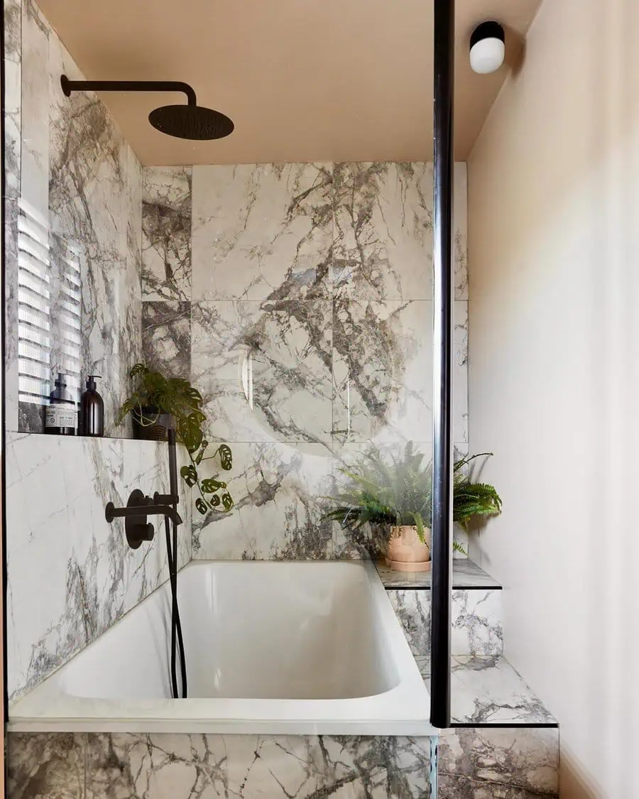 Small luxury bathroom with marble walls, black fixtures, plants, and a built-in bathtub.