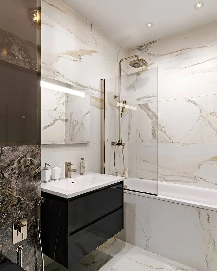 Small luxury bathroom with marble walls, black vanity, glass shower, gold fixtures, and luxe lighting.