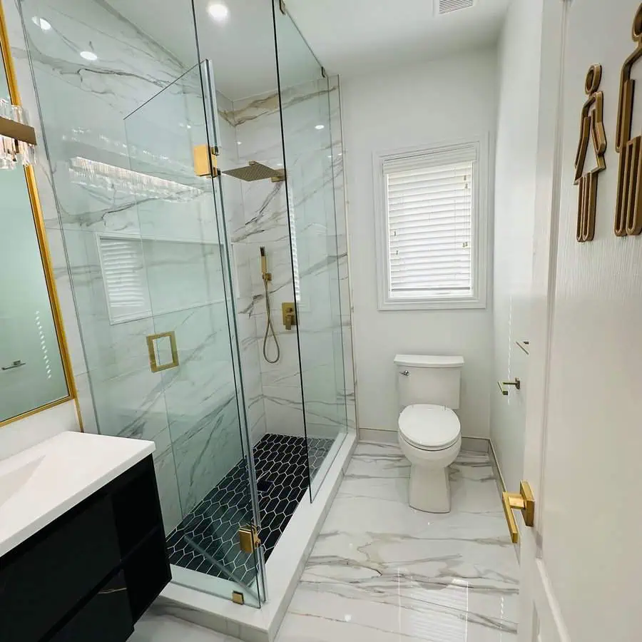 Small luxury bathroom with marble tiles, gold accents, glass shower, and black vanity.